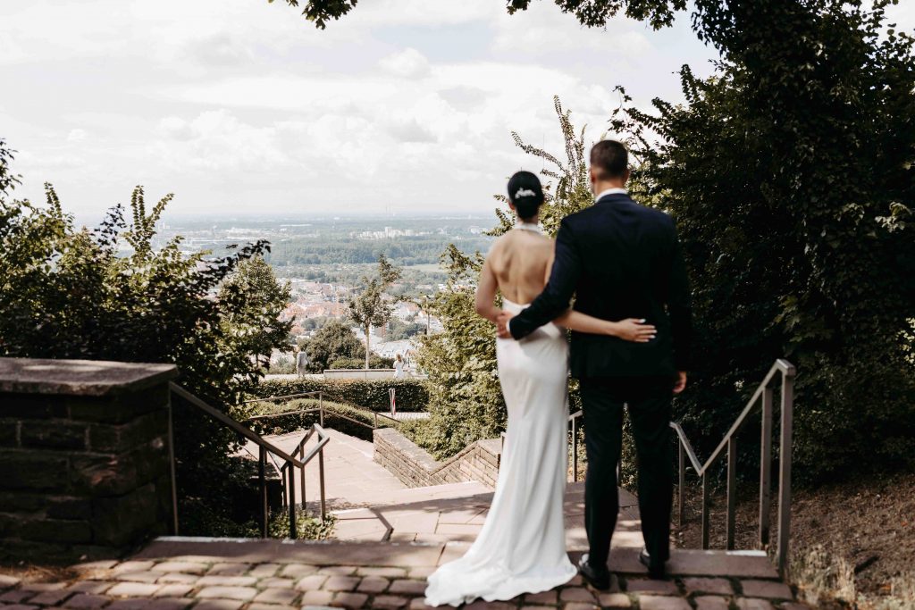 Das Brautpaar umarmt sich hinterm Rücken. Beide schauen über die Stadt Karlsruhe.