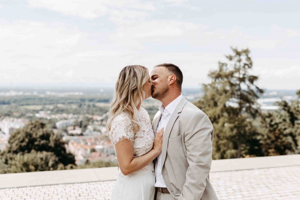 Ein sich küssendes Brautpaar. Die Braut legt ihre Hand mit dem Ehering liebevoll auf seine Brust. Er hat lässig seine Gang in der Hosentasche. Beide stehen vor dem Standesamt auf dem Turmberg in Karlsruhe. Man sieht im Hintergrund den Horizont. So weit kann man über die Stadt Karlsruhe blicken.