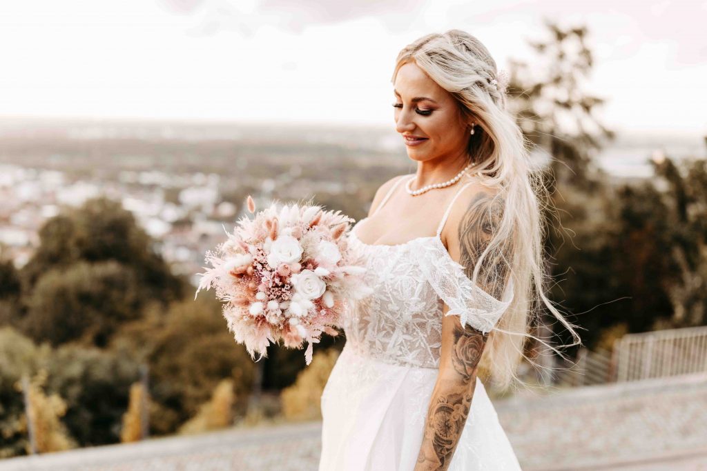 Hochzeitsfotograf Karlsruhe: Die Braut schaut ihren Brautstrauß an. Eine leichte Brise weht durch ihr Haar. Der Sonnenuntergang auf dem Turmberg Karlsruhe ist zu spüren. Im Hintergrund sieht man den Weitblick über die Stadt Karlsruhe
