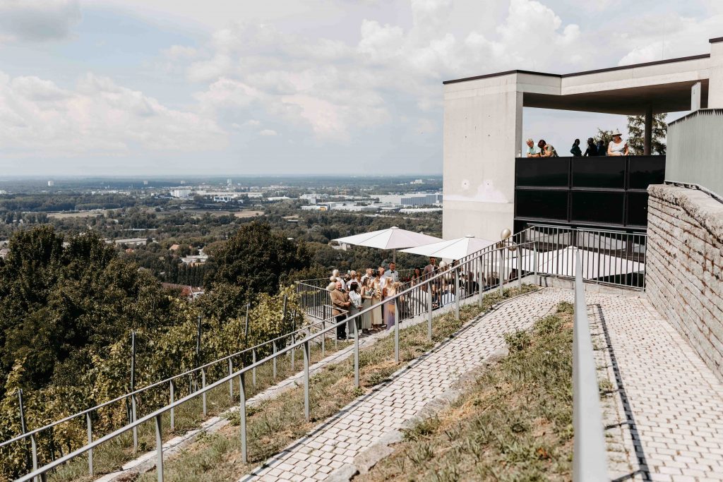man sieht den Weitblick über Karlsruhe vom Standesamt Durlach auf dem Turmberg aus. Man sieht teilweise das Gebäude, den Trausaal sowie die Hochzeitsgesellschaft auf der Terrasse