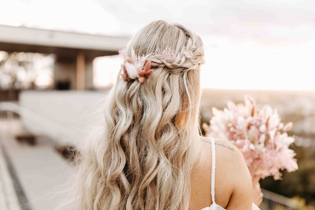 man sieht eine Braut von hinten, die ihren Brautstrauß anschaut. Der Fokus liegt auf der Frisur der Braut. Im Hintergrund sieht man den Weitblick über Karlsruhe. Das Bild entstand auf dem Turmberg Karlsruhe