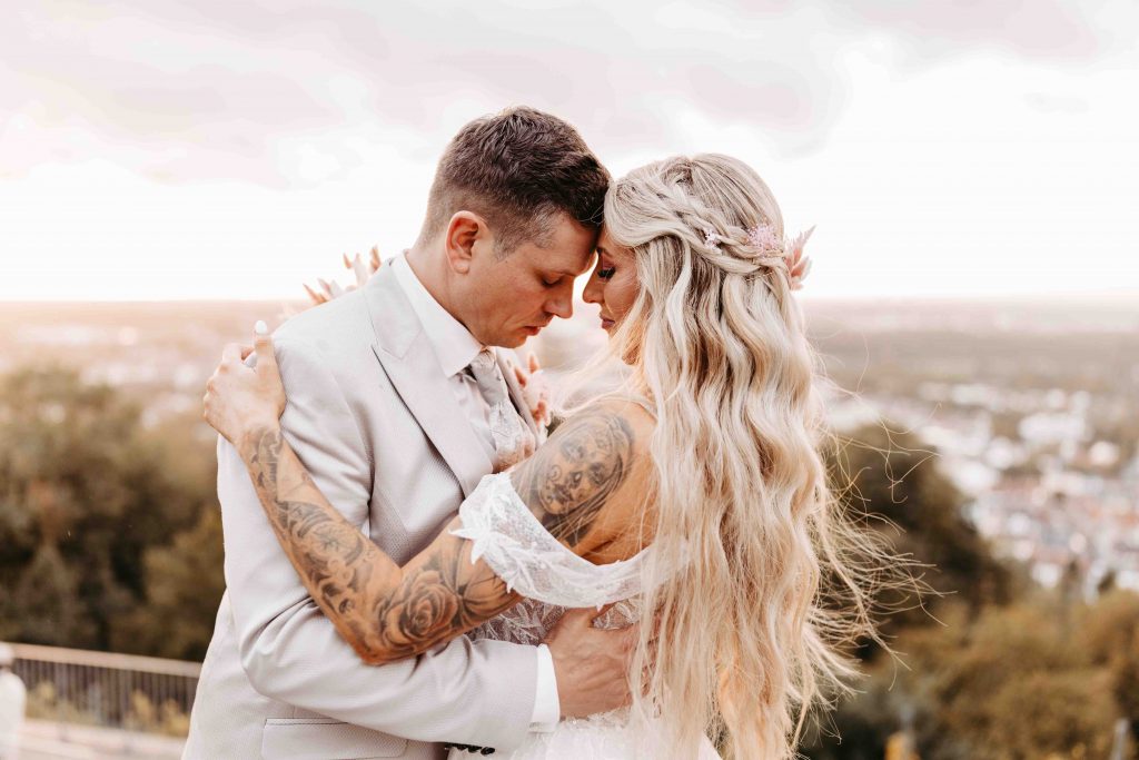 Verliebtes Brautpaar. Beide legen ihre Stirn aneinander. Sonnenuntergangsstimmung. Die Augen des Paares sind geschlossen. Sie umarmen sich liebevoll. Man sieht im Hintergrund den Weitblick über die Stadt Karlsruhe
