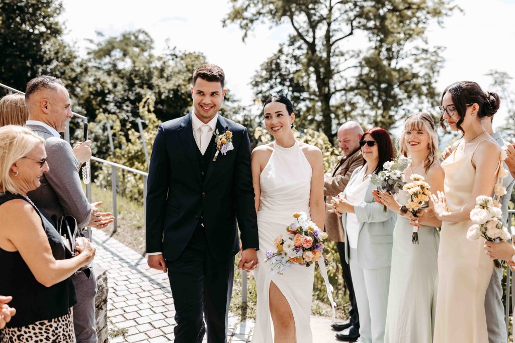 Das Brautpaar wird von der Hochzeitsgesellschaft empfangen. Die Gäste klatschen. Das Paar hält sich an den Händen und geht durch die Mitte durch in Richtung Trauung