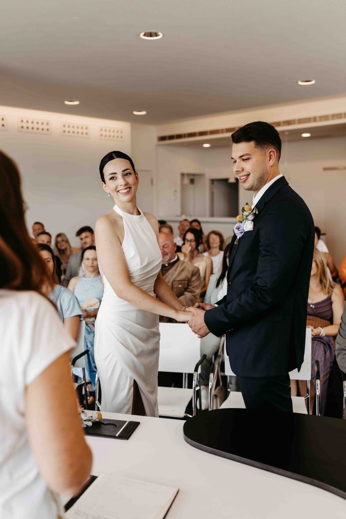 Während der Trau-Zeremonie im Trausaal auf dem Turmberg in Karlsruhe. Das Brautpaar hält sich and den Händen und schauen die Standesbeamtin an.