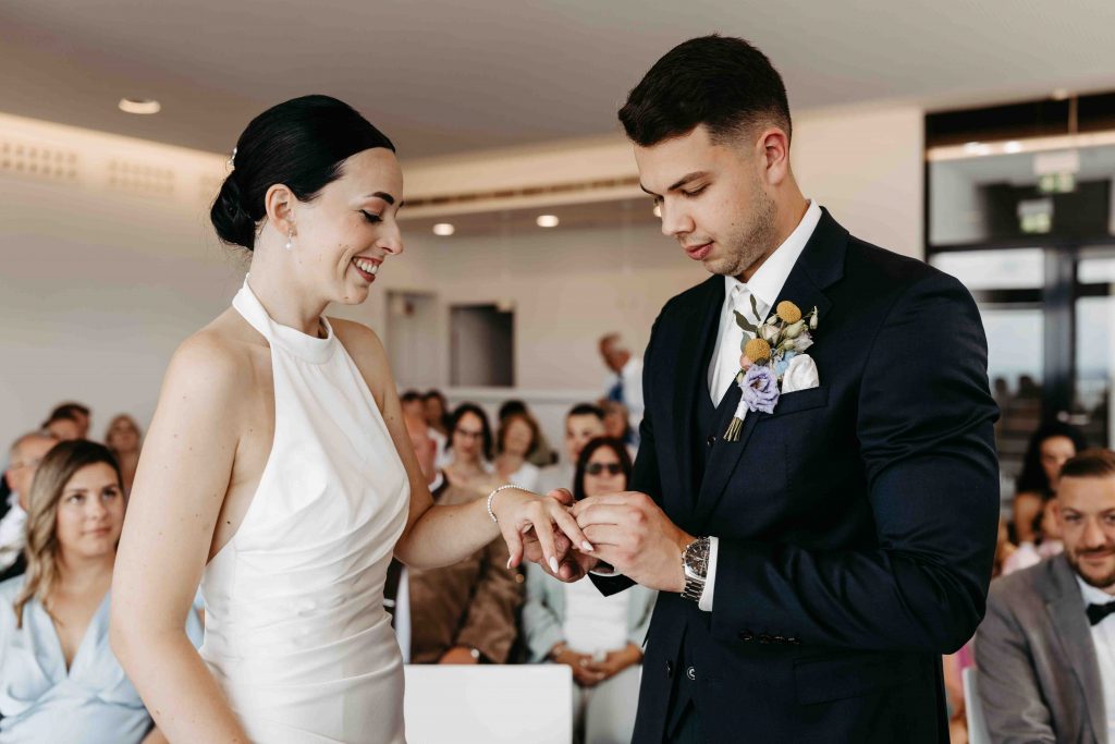 Ringtausch während der Trau-Zeremonie. Der Bräutigam steckt der Braut den Ring an den Finger. Sie schauen beide auf die Hand der Braut und sie lächelt und strahlt.