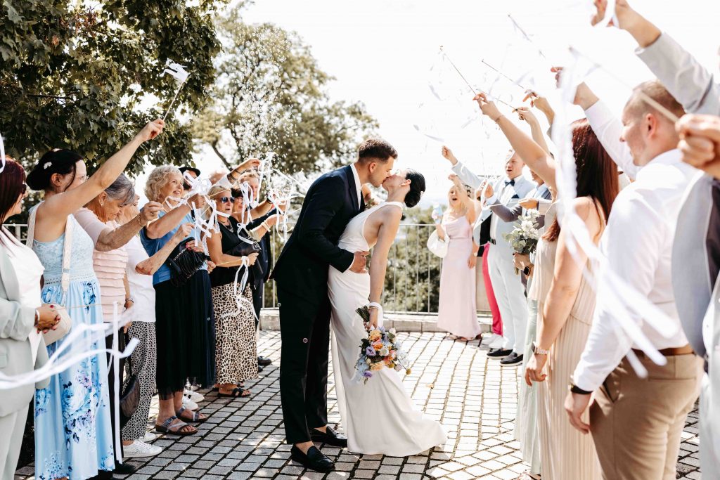 Ein Brautpaar steht inmitten eines Spaliers. Die Gäste pusten Seifenblasen und jubeln mit Bändern an kurzen Stöckchen. Das Paar küsst sich romantisch. Die Braut geht dabei ins Hohlkreuz