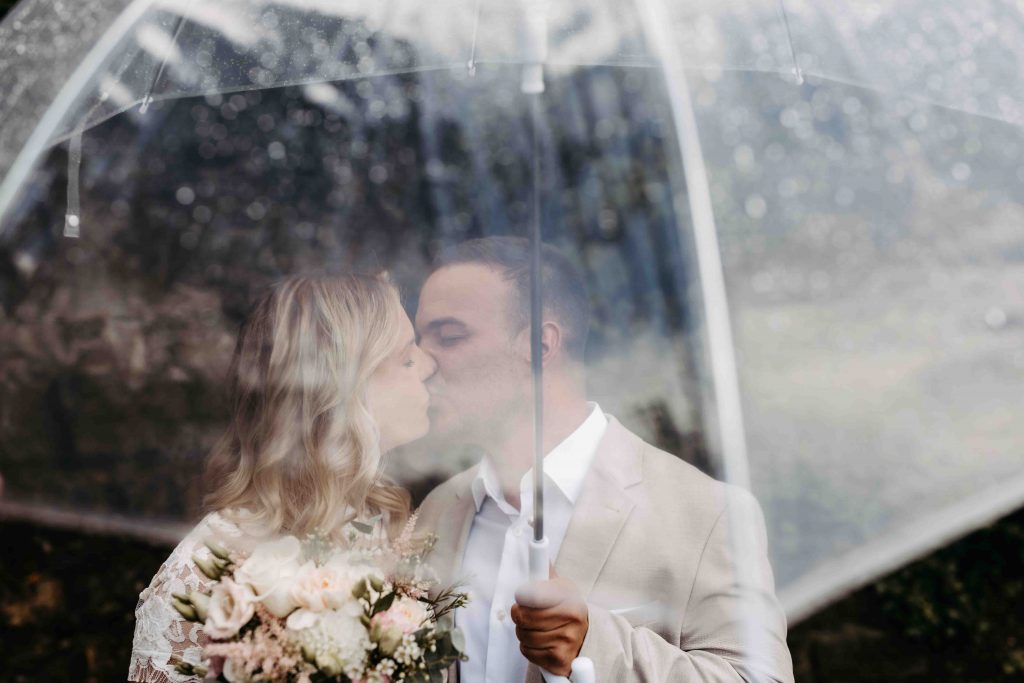 Das Brautpaar steht unter einem transparenten Regenschirm. Die Braut hält ihren Brautstrauß und erhält den Schirm. Das Foto ist so gestaltet, dass durch den Schirm hindurch fotografiert wurde. Das Paar küsst sich