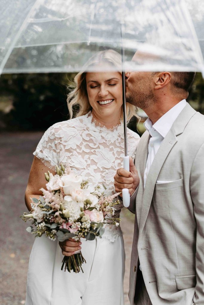 Das Brautpaar steht unter einem Regenschirm. Er gibt ihr einen Kuss auf die Schläfe. Sie genießt den Moment und schließt ihre Augen. Sie lächelt.