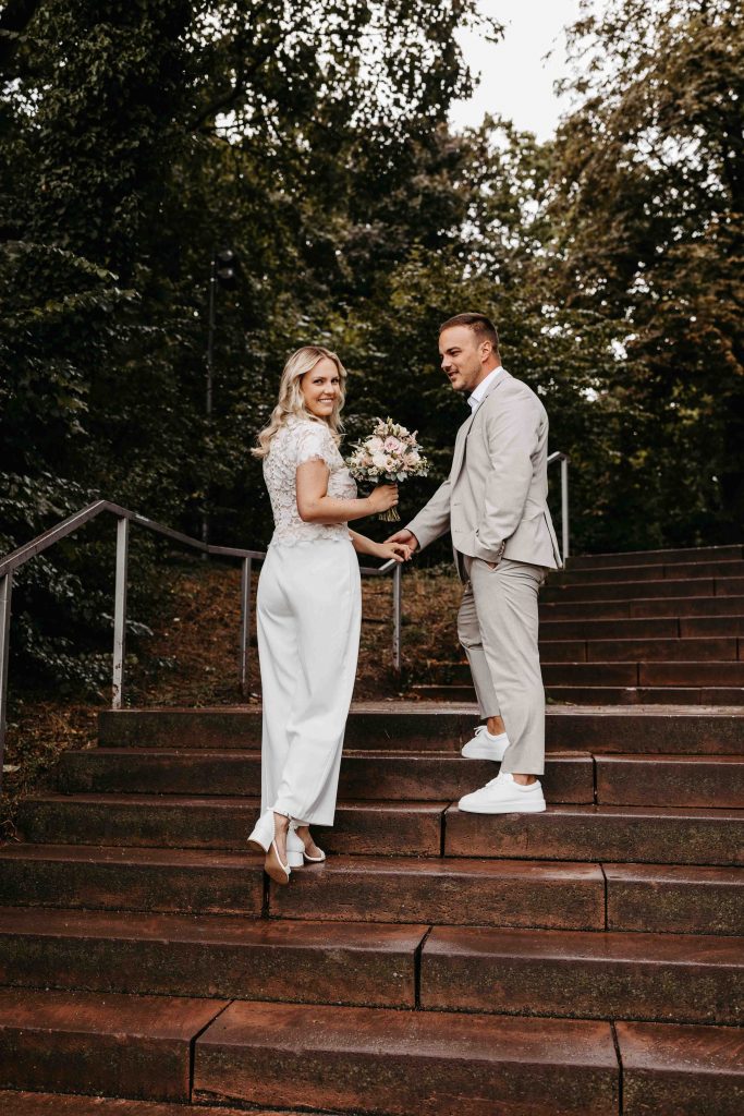 Der Bräutigam hält die Hand der Braut während sie die Treppen zum Park hinaufgeht. Sie wirft ihren Blick in die Kamera. Er schaut sie an.