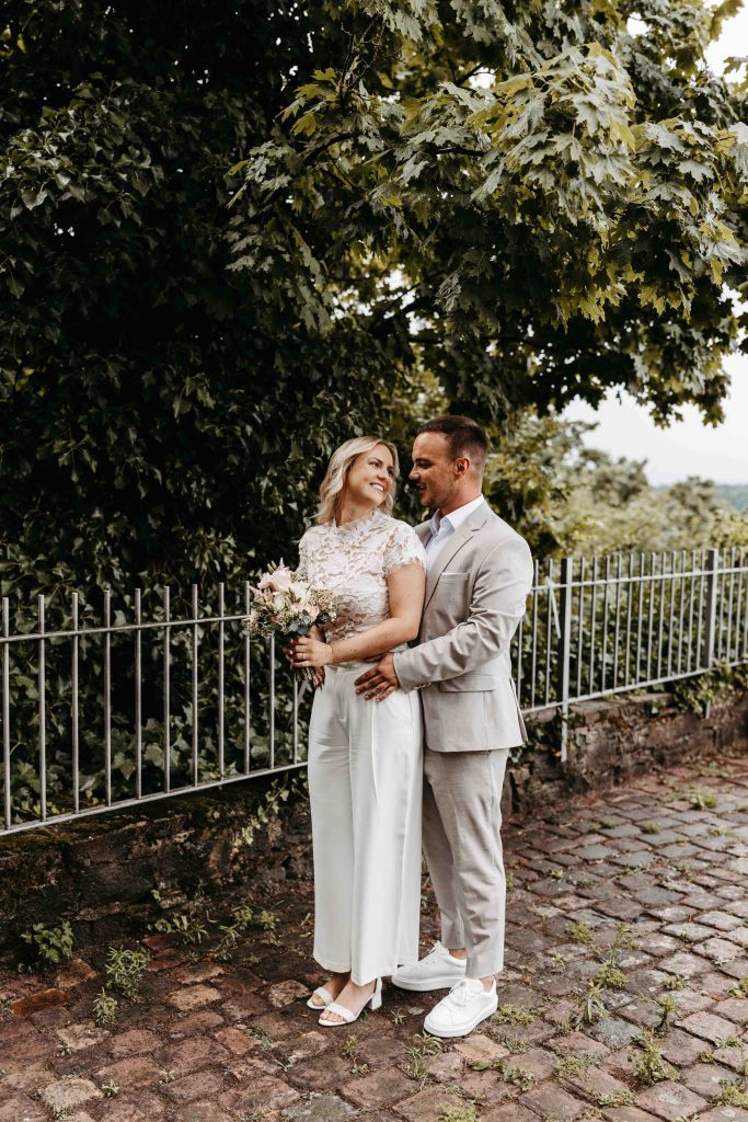 Das Brautpaar steht unter einem Baum, nahe des Aussichtspunkt auf dem Turmberg in Karlsruhe-Durlach. Er steht hinter ihr, seitlich, und sie lehnt ihren Rücken an seine Brust. Sie dreht den Kopf zu ihm und lächelt ihn an.