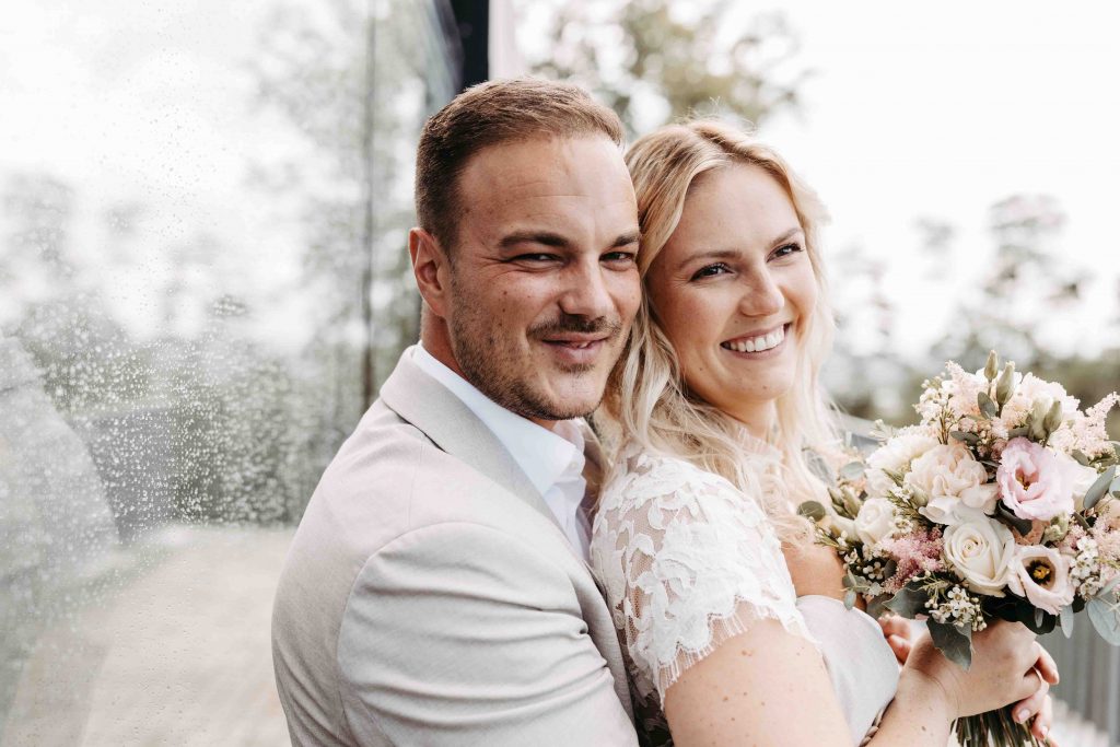 Das Brautpaar lächelt herzlich. Sie ist mit dem Rücken an ihn angelegt. Er umarmt sie und schaut dabei in die Kamera. Sie schaut in die Ferne und hält dabei ihren Brautstrauß in den Händen