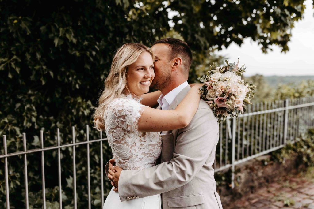 Brautpaar ist umschlungen. Er küsst sie auf die Schläfe, sie strahlt seitlich in die Kamera mit Blick in die Ferne. Sie umarmt ihn hinter seinem Nacken und hält dabei ihren Brautstrauß in den Händen. Er umarmt ihre Hüfte