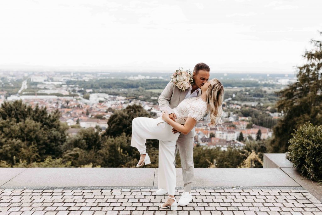 Das Brautpaar küsst sich. Dip-Kiss. Das Paar steht mittig im Bild. Drumherum ist ein Panorama-Blick über die Stadt Karlsruhe