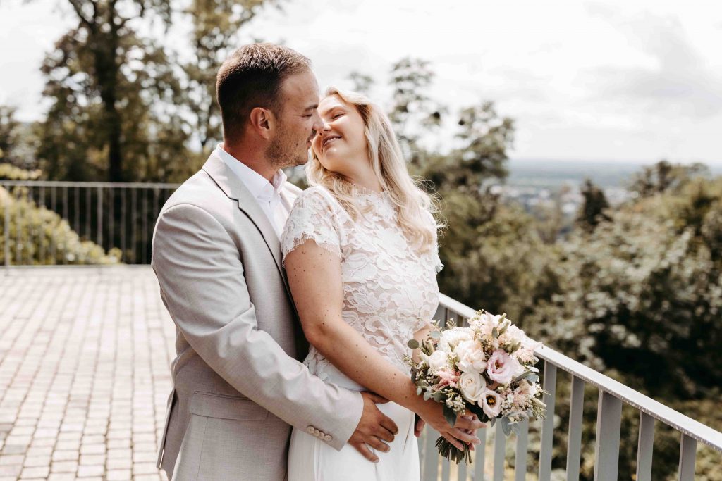 Das Brautpaar steht am Geländer der Terrasse des Trausaals Turmberg in Durlach. Er umarmt seine Braut von hinten. Sie hält ihren Brautstrauß in den Händen und dreht ihren Kopf zu ihm nach hinten, um ihn zu küssen.