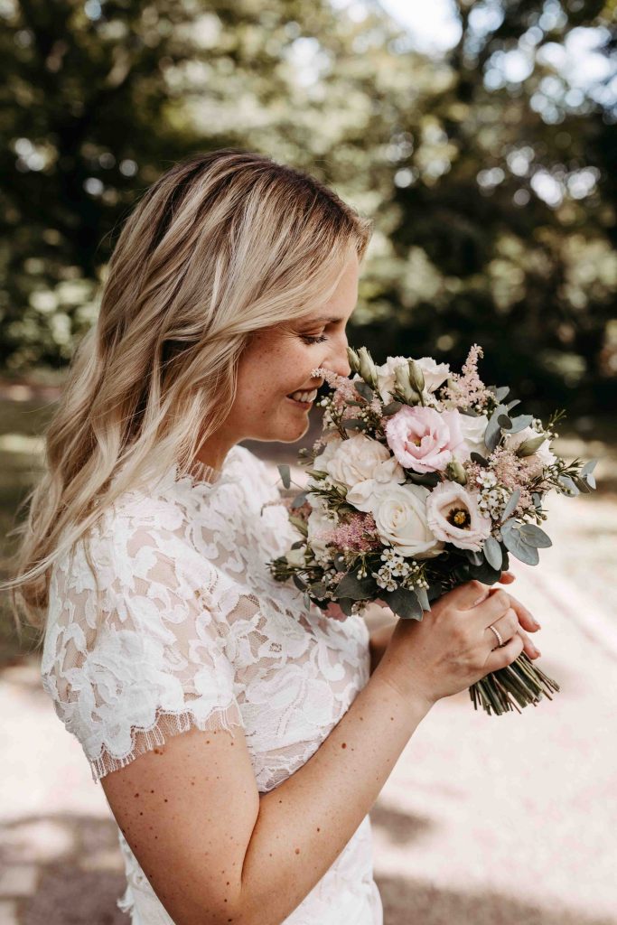 Die Braut riecht an ihrem Brautstrauß. Sie lächelt glücklich und schaut den Strauß dabei an. Sie hält ihn in ihren Händen.