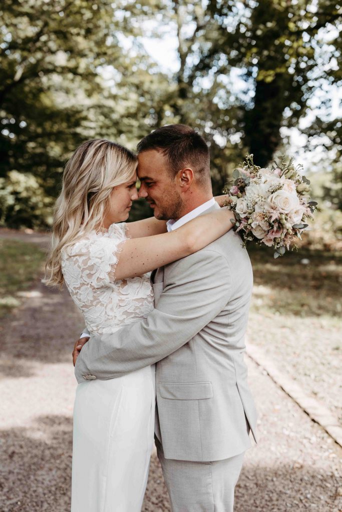 Das Brautpaar schaut sich tief in die Augen. Stirn an Stirn. Sie umarmt ihn, in dem sie ihre Arme um seinen Nacken legt. In den Händen hält sie ihren Brautstrauß. Er umarmt sich an der Hüfte