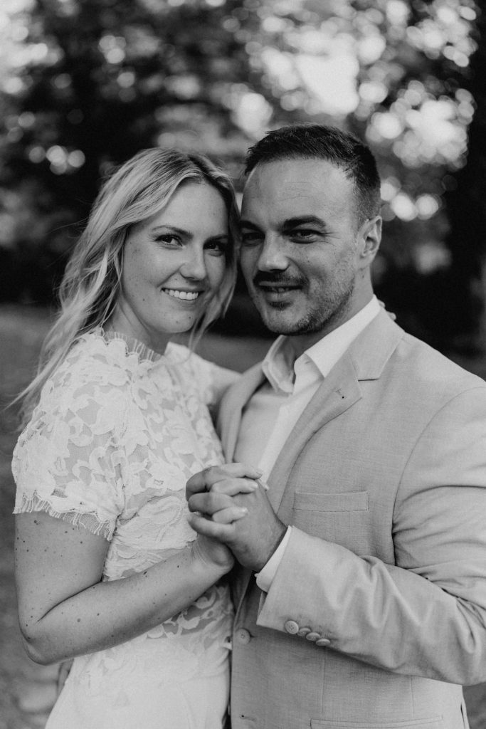 Das Brautpaar schaut direkt in die Kamera. Ihre Haltung ist, als würden sie gerade tanzen. Ihre Köpfe haben sie aneinander gelegt.