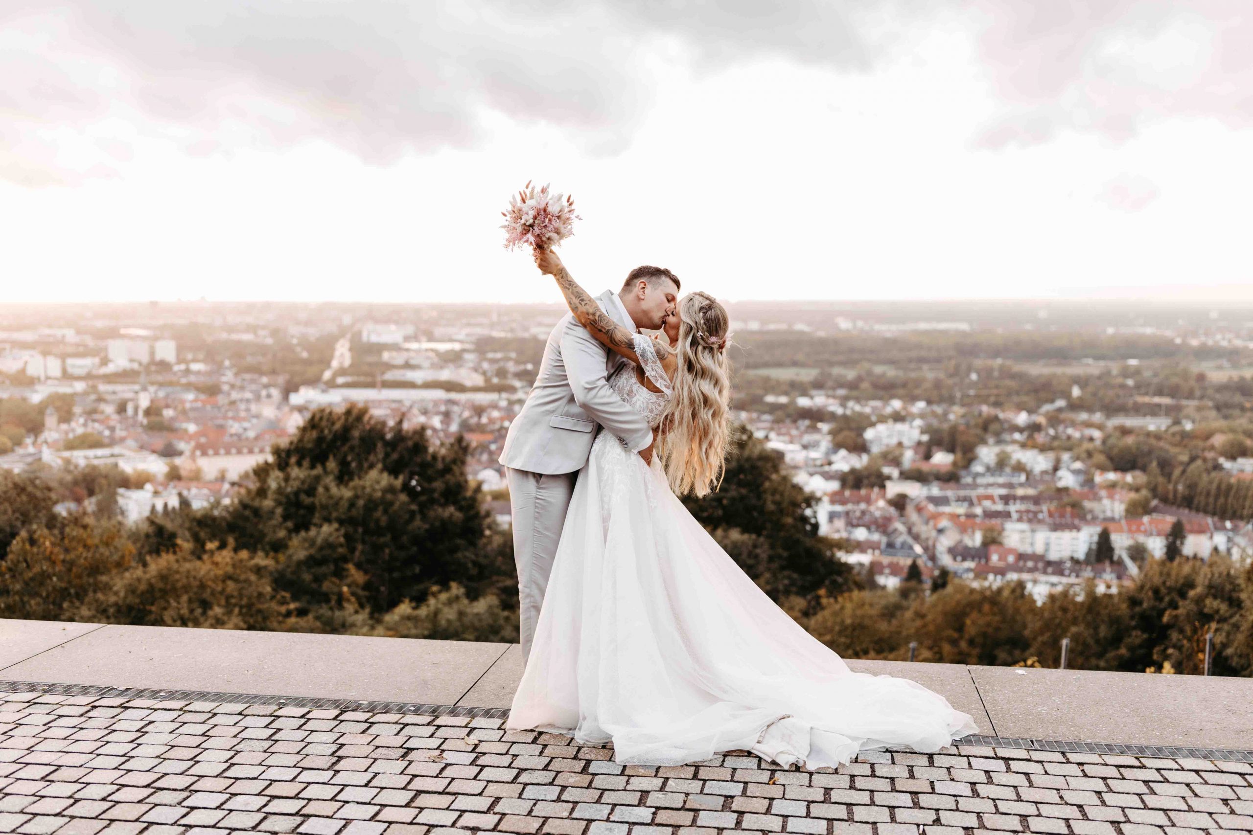 Hochzeitsfotograf Karlsruhe: Das Brautpaar steht auf dem Turmberg in Karlsruhe-Durlach. Das Brautpaar steht in der Mitte und küsst sich. Sie hält ihren Brautstrauß in die Höhe und geht dabei leicht ins Hohlkreuz. Sie stehen mittig. Sie sind umgeben von einem Panorama-Blick über die Stadt Karlsruhe