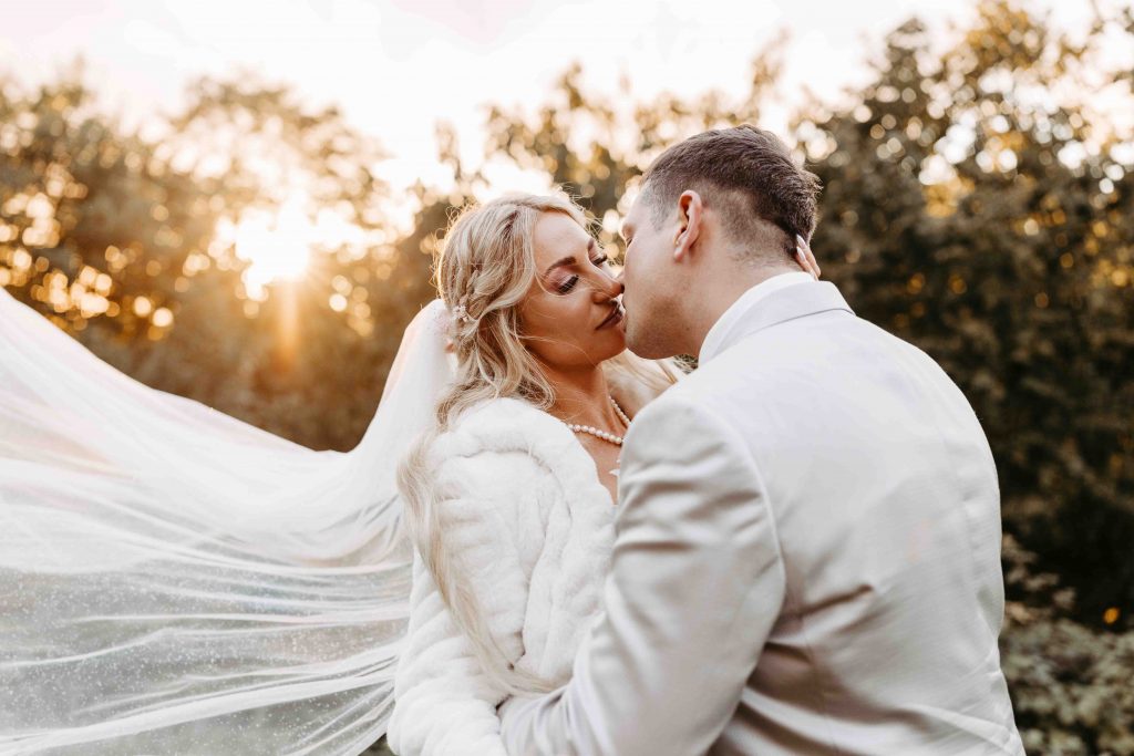 ein inniges Foto. Das Brautpaar ist kurz vor dem Kuss. Die Sonne geht im Hintergrund unter und funkelt durch die Bäume im Hintergrund. Das Brautpaar hat die Augen geschlossen. Der Wind weht durch den Schleier.