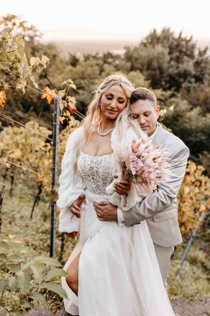 Das Brautpaar steht zwischen den Weinreben. Es ist Herbst. Der Bräutigam umarmt seine Braut von hinten und beide haben die Augen geschlossen und genießen den Moment.