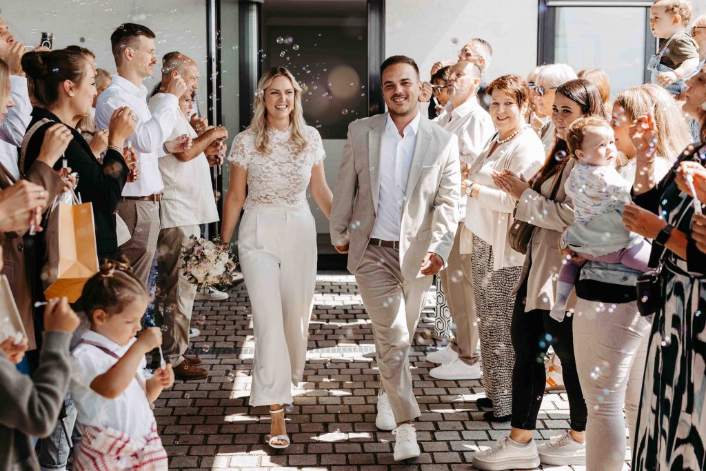 Das Brautpaar, frisch vermählt, läuft aus dem Standesamt. Ein Spalier hat sich gebildet, die Leute pusten Seifenblasen auf das Brautpaar.